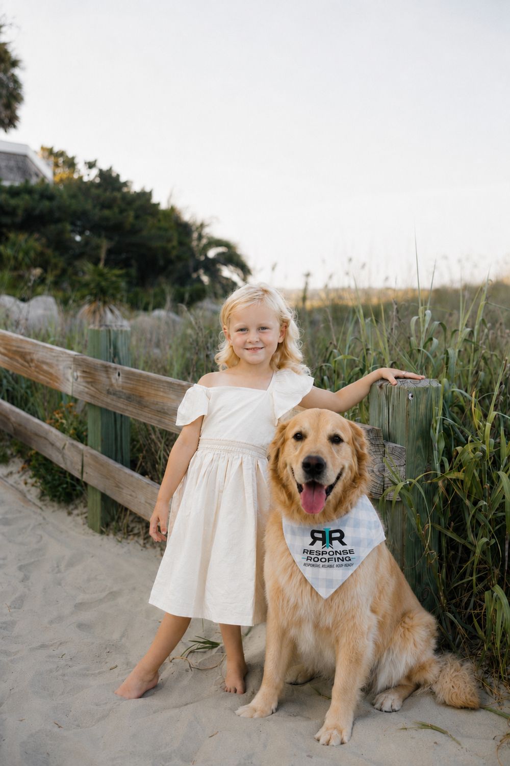 A young girl and a golden retriever wearing a Response Roofing bandana on a South Carolina beach walkover at sunset.